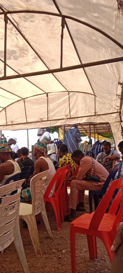 This image depicts an outdoor gathering in Iyara, Nigeria, under a large, temporary tent structure constructed from metal poles and a white tarp roof. The ground beneath the tent is reddish-brown earth with small stones.
Approximately twelve to fifteen individuals are visible, predominantly male, seated on plastic chairs of various colors including white, red, and blue. In the foreground, from left to right, a person in a white tank top and a patterned traditional hat is seated on a white chair, facing right. Next to them, another individual wearing a white tank top and a similar patterned hat is also seated on a white chair, viewed from the side. A third individual is seated on a red chair, wearing a light brown or peach-colored long-sleeved shirt and trousers, looking downwards. This person has a multi-colored strap across their chest and a small dark bag hanging from it.
In the mid-ground, other attendees are visible. One individual wears a yellow and black patterned shirt. Further back, a person is seen from behind, wearing a white and blue striped traditional garment. Another person wears a white t-shirt. Most individuals are seated, some appear to be observing, while others, like the person in peach-colored clothing, are looking down, possibly at a device not fully visible. The tent provides shade, and the lighting is even.