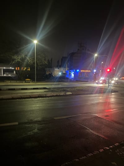 The image depicts a nighttime street scene in Schweinfurt, Germany, likely after rain due to the wet, reflective asphalt road. The perspective is from the roadside, looking down a street.

In the foreground, a dark, wet asphalt road stretches into the distance, marked by faint white lines and reflecting the bright artificial lights. To the right, a dark curb borders the road.

Along the left side of the road, multiple streetlights emit bright, starburst-like white light, illuminating the bare branches of a tree and parts of the dark surroundings. Further back on this side, a structure resembling a bus stop or shelter is visible, and behind it, a large, dark building with some flags nearby.

On the right side of the road, a prominent building is brightly lit with distinctive blue and orange lights. The German word "SPIELHALLE" (meaning "arcade" or "gaming hall") is clearly visible in orange letters on a blue background on this building. In front of this building, several vehicles are present, their headlights and taillights creating streaks of white and red light. A red traffic light is also visible in this area. The sky above is dark, confirming the nighttime setting. No people are explicitly visible, and the scene captures a functional urban environment at night.