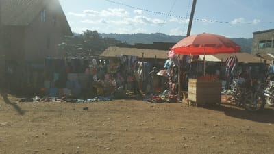 The scene is a small marketplace in Kayna, Democratic Republic of the Congo. A red umbrella shades a stall displaying various colorful clothing items on a wall behind the stall. There are several people around the stall, some looking at merchandise.  A wooden crate sits in front of the stall containing what appears to be oranges. A motorcycle is parked to the right of the stall.  The ground is dirt, and in the distance, a hillside with scattered trees and vegetation can be seen. The sky is partly cloudy.  The surrounding buildings are mostly made of brick.  The scene is bright and sunny.