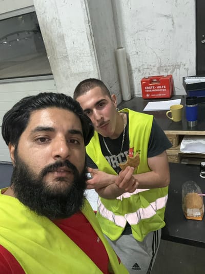 Two men are in a dimly lit, industrial-looking room. The man in the foreground, facing the camera, has dark hair and a beard, and is wearing a yellow safety vest over a red shirt. He is looking directly at the camera, with his face close to the lens. Behind him, another man, with short hair and a mustache, is looking to his right and holding a waffle cookie in his left hand. He is wearing a similar yellow safety vest over a dark t-shirt and a silver chain. In the background, there is a first-aid kit on a shelf, a yellow mug, a blue thermos, and some rolls of material. The walls appear to be concrete. There is also a plastic-wrapped item, possibly food, on a table in the right foreground. The scene suggests a break or casual moment at a workplace.