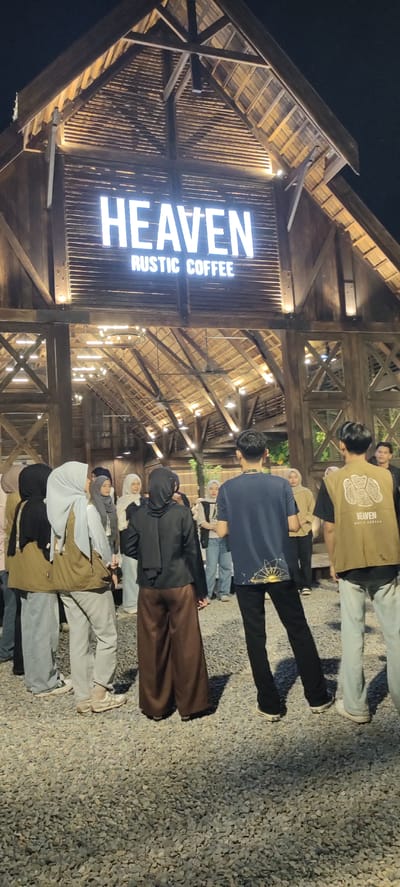 A group of people stand outside a rustic coffee shop called "HEAVEN RUSTIC COFFEE" at night in Bireuenmeunasahdayah, Indonesia. The building has a wooden exterior and a wooden roof with exposed beams and rafters. The name of the coffee shop is illuminated with white neon letters. The ground is covered in gravel. In the foreground, several individuals are visible, some facing away from the camera. One person in the center foreground wears a dark blue t-shirt and black pants. To their right, another person wears a brown vest with the coffee shop's logo and light blue jeans. Several women in the middle ground wear hijabs and various colored clothing, including a black hijab and brown pants on one individual, and a light blue hijab and grey pants on another. The scene suggests a social gathering at an outdoor establishment.