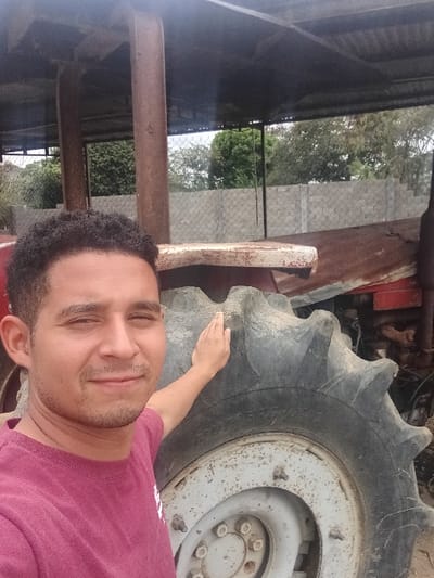 A male individual with short dark curly hair and light facial stubble, wearing a maroon t-shirt, smiles directly at the camera. His right hand rests on the deeply-treaded, dirty black rubber tire of an old, red-painted tractor exhibiting extensive rust; the tractor's light gray wheel hub is also visible. The scene is set under a corrugated metal roof supported by thick, rusted reddish-brown metal posts. In the background, beyond the structure, a chain-link fence, a concrete wall, and dense green foliage are visible under daylight conditions. The location is Cujisal, Venezuela.