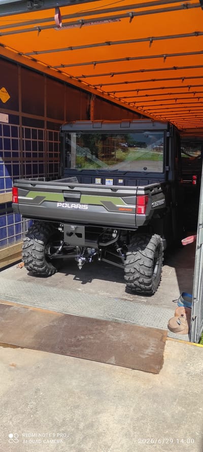 In the city of Valga, Estonia, a dark grey Polaris Ranger utility vehicle is parked inside a truck or trailer with an orange tarp-like roof. The vehicle is seen from the rear, with its large, knobby tires prominently displayed. The bed of the vehicle features a green and grey graphic design and the "POLARIS" and "RANGER" logos. The rear window of the cab is visible, reflecting an outdoor scene. The truck or trailer interior has metal support beams and corrugated metal walls in some areas. Straps and some boots are visible near the entrance of the trailer. The ground outside the trailer appears to be concrete or asphalt, with some metal plating near the trailer's ramp. A watermark in the lower left corner indicates the photo was taken with a Redmi Note 9 Pro and AI Quad Camera, and the timestamp reads "2026/6/29 14:00".