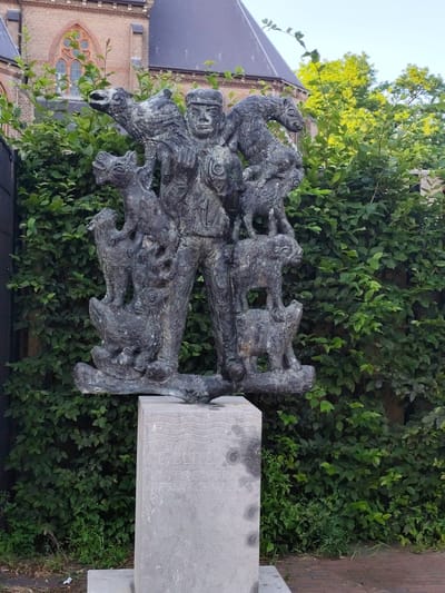 This image shows a bronze statue of a man surrounded by various animals, including dogs, sheep, and birds. The man appears to be a shepherd or farmer, with a hat on his head and rugged clothing. The animals are depicted in different positions, some on his body, and others around him. The statue is mounted on a light grey stone pedestal, on which some text is engraved, though it is partially obscured and difficult to read in full. The statue and pedestal are set against a backdrop of lush green bushes and trees. In the background, a brick building with a dark roof and a gothic-style window is visible. The lighting suggests it is daytime, possibly late afternoon, with sunlight filtering through the trees and casting shadows. The overall impression is one of a historical or allegorical representation of a person connected to nature and animals, possibly in a rural setting. The location context provided is Schaijk, Netherlands.