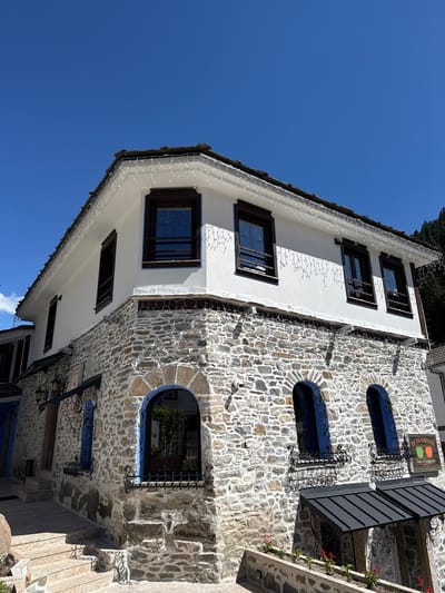 A two-story building with a stone-clad lower level and a white-painted upper level is depicted in Shiroka Laka, Bulgaria. The building has a tiled roof and is adorned with white string lights along the eaves. The lower level features arched windows with blue trim and decorative metal grates. The upper level has rectangular wooden-framed windows. A sign reading "YA TA VIDVAM Optics & Sunglasses" is visible on the right side of the building. The scene is set against a clear blue sky, and a forested hillside is visible in the background. There are no people or animals present in the image.