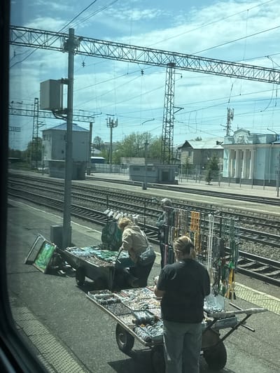 The image depicts a scene at a railway station in Kropachyovo, Russia, as viewed from inside a train. The view is across the railway tracks. There are at least two individuals near a makeshift jewelry stand or cart. The cart holds a variety of jewelry items. The background consists of railway infrastructure, including tracks, electrical lines, support structures, and buildings typical of a railway environment. The weather appears to be sunny with some clouds in the sky.