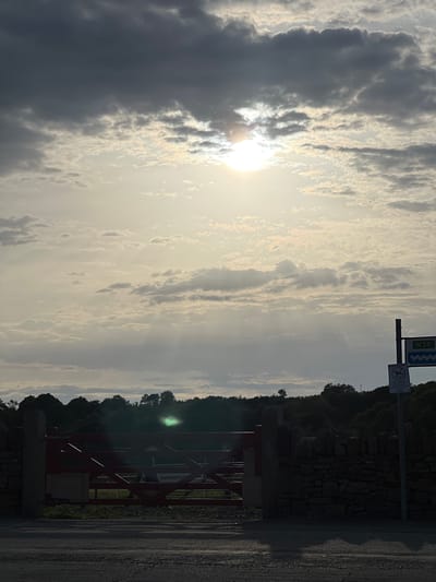 A view of a red wooden gate and a stone wall with a road in the foreground. In the background, a treeline is visible under a cloudy sky with the sun shining through, casting sunbeams. There is a road sign visible to the right of the image. The scene is located in Terhillion, Ireland.