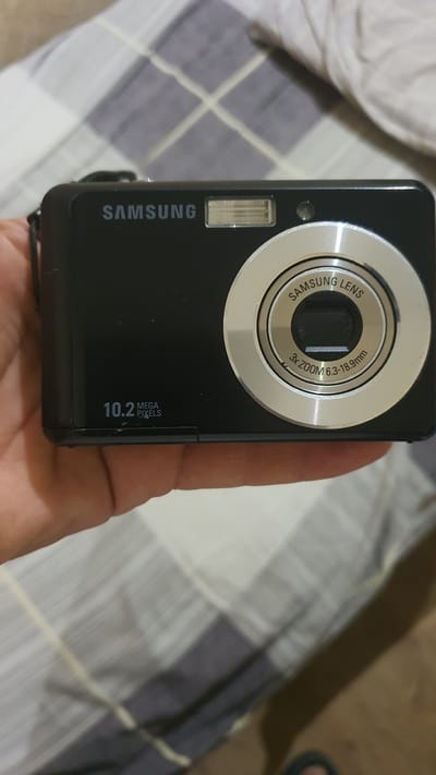 A black Samsung digital camera is held in a person's hand, with the camera's lens and branding prominently displayed. The camera has "SAMSUNG" written in silver text above the flash unit, and "10.2 MEGA PIXELS" is visible on the lower left side of the camera body. The lens assembly features "SAMSUNG LENS" in a circular engraving, along with "3x ZOOM 6.3-18.9mm". The background consists of a bed with a checkered duvet cover in shades of grey and white. No specific location within the city of Karankul, Uzbekistan is identifiable from the image.
