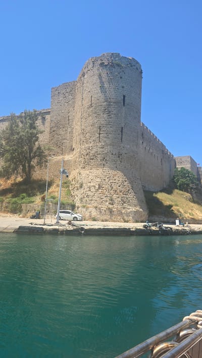 This is a vertical, eye-level, full-shot photograph of Kyrenia Castle in Kyrenia, Northern Cyprus. The castle is a large, imposing stone structure with a prominent round tower on the left side. It's situated on a hill overlooking a body of water that takes up the bottom third of the frame. The water is a vibrant turquoise color with gentle ripples.

Along the edge of the water, a concrete walkway runs in front of the castle walls. A white car is parked on this walkway, near a lamppost with a security camera attached. There are also various ropes, buoys, and small boats docked along the edge.

The castle walls are made of large, rough-hewn stones and show signs of age, with some vegetation growing on the ramparts. The sky above is a clear, bright blue. The overall scene suggests a sunny day in a historic coastal city.