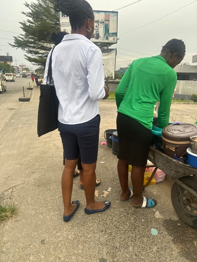 Two women stand beside a cart filled with cooking supplies. One woman, in a green shirt and black skirt, faces the cart. The other woman, in a white shirt and navy shorts, faces away from the cart and towards a busy street. She carries a black bag. The cart appears to have pots and buckets. They both wear sandals or flats. The background shows a street scene with a billboard, trees, and passing cars.