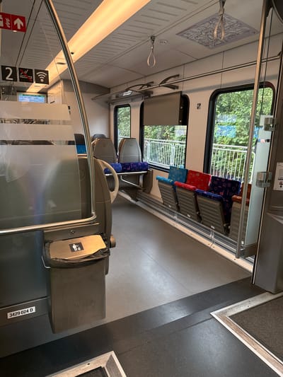 The image depicts the interior of a passenger train carriage in Velbert, Germany. The perspective is from a low angle, looking forward down the aisle. The carriage features rows of seats with blue and red patterned upholstery, and some seats are facing forward while others are facing backward. Large windows line the right side of the carriage, offering a view of green trees and foliage outside.

In the foreground, on the left side, a large, metallic trash receptacle is visible. Above it, a frosted glass partition is in place. Further to the left, a digital display screen shows route information, indicating "S9 Velbert am See" and "16:22 Velbert-Nierenhof". There are also signs visible on the wall, including a large number "2" and symbols indicating no smoking and Wi-Fi availability.

The ceiling of the carriage is illuminated by long, fluorescent light fixtures. Several hand straps are hanging from the ceiling, and a map is affixed to the ceiling above the seats. The floor is a combination of gray and black textured material. A metal railing runs along the right side of the aisle. The overall impression is one of a clean and modern train interior.