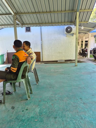 Scene in Pulorungkom, Indonesia, under a covered structure with a corrugated metal roof. Two men in work uniforms are interacting closely. One man is seated in a green plastic chair, wearing a green uniform with orange stripes and an orange safety vest. The second man, dressed in a yellow shirt, stands behind him, with his hand on the seated man's back. A white wall is behind them, with an air conditioning unit mounted. Motorbikes are visible in the background, parked on a paved area. The floor is painted in a teal color, showing wear.
