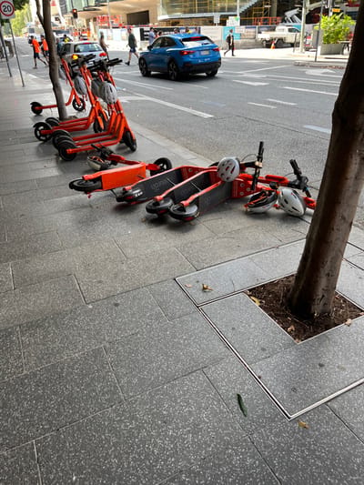 The image captures a vibrant urban street scene, likely a sidewalk area, populated with a row of parked electric scooters. The perspective is slightly elevated, looking down along the length of the street.

In the foreground and mid-ground, a cluster of orange Neuron electric scooters is visible, some neatly lined up, others a bit askew. Each scooter has a white helmet resting on its handlebars or seat. The brand name "Neuron" is prominently displayed in white text on the footboard of the scooters. These scooters are parked alongside a large tree trunk that emerges from a square metal grate set into the grey, speckled concrete pavement. Further along the sidewalk, more scooters of the same model are visible.

To the left of the scooters, the paved street is visible, with white road markings. A blue SUV is driving away from the viewer, and further down the street, another white vehicle is parked. Several pedestrians are walking on the sidewalk and crossing the street in the background, suggesting a moderately busy environment. One pedestrian is visible near the blue SUV, and another further back.

The setting appears to be daytime, with soft, ambient lighting suggesting an overcast day or perhaps early morning/late afternoon. The general ambiance is functional and utilitarian. A small, dark green elongated object (possibly a leaf or debris) is visible on the pavement near the tree grate in the mid-right. In the bottom-left corner, a round white sign with the number "20" is partially visible, indicating a speed limit of 20 km/h, which is common in urban areas, including South Brisbane, Australia. The architecture in the background consists of modern buildings, contributing to the urban feel.