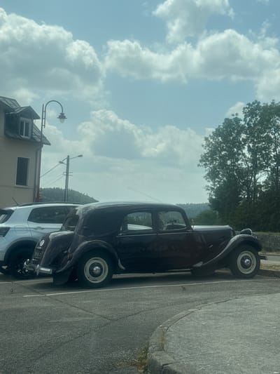 In La Chaux-du-Dombief, France, a dark maroon vintage Citroën car is parked next to a modern white SUV in a paved parking lot. The Citroën, with its distinctive curved fenders and elegant silhouette, is positioned in the foreground. The white SUV is slightly behind and to the left of the vintage car. In the background, a building with a peaked roof and a window is visible to the left, and a streetlamp stands nearby. Further back, a line of trees and a hillside create a natural backdrop under a sky filled with white, fluffy clouds. The scene appears to be a still, outdoor environment during daylight.