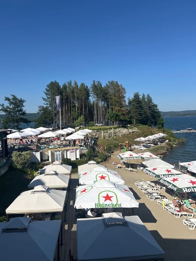 The image shows a sun-drenched beach resort in Dolni Pasarel, Bulgaria. White and green Heineken-branded umbrellas are scattered across the sandy beach, providing shade for numerous lounge chairs occupied by people sunbathing and relaxing. A wooden boardwalk separates some of the seating areas from the sand. In the background, a calm body of water, likely a lake, is visible, with a small boat docked at a pier. Lush green trees and a dense forest line the edge of the water, creating a picturesque backdrop. The sky is clear and blue, indicating a bright, sunny day.