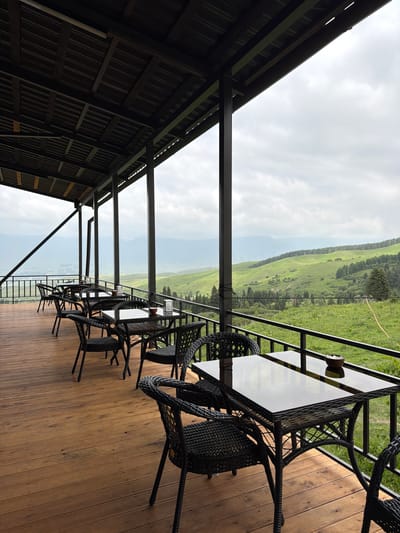 The image shows an outdoor covered patio with multiple tables and chairs, overlooking a lush green mountainous landscape. The patio has a wooden floor and a dark, slatted roof supported by vertical beams. Black wicker tables with reflective dark tabletops are set up, each with several matching wicker chairs. Some tables have small bowls on them. A black metal railing runs along the edge of the patio, beyond which the scenic view unfolds. The landscape consists of rolling hills covered in green grass and dotted with trees, with mountains visible in the distance under a cloudy sky. The scene is serene and appears to be a restaurant or cafe setting. It is located in the city of Kaindy, Kyrgyzstan. There are no people visible in the image.