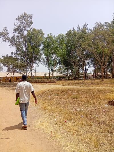 An outdoor scene in Ban, Nigeria, depicts a lone male subject walking away from the camera along an unpaved, reddish-brown dirt path. The subject, a dark-skinned male, wears a light-colored short-sleeved collared shirt, blue denim jeans, and brown sandals, carrying a bright green folder or book in his left hand. To the right of the path, the ground is covered in dry, yellowed grass and scattered litter, including plastic containers. The background features several low-rise buildings with light-colored walls and reddish roofs, partially obscured by numerous trees with varying foliage density, some lush green, others sparser. The sky above is a pale, bright blue. The environment suggests a dry, possibly arid, season and an institutional or campus-like setting.