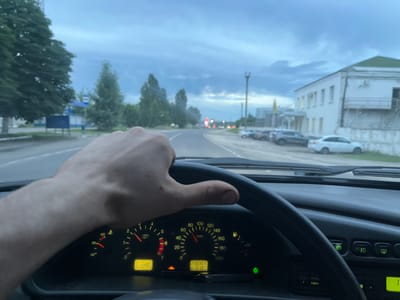 The image is a first-person perspective from inside a car, looking out through the windshield. The driver's left hand is on the steering wheel. The dashboard shows the speedometer indicating approximately 60 km/h, the tachometer around 1500 rpm, the engine temperature gauge at 90°C, and the odometer reading 230307 km. The fuel gauge is slightly above 1/2. Outside, it appears to be dusk with cloudy skies. The car is on a paved road with trees and buildings lining the sides. There is a bus stop with signage on the left side of the road, and several parked cars on the right. A white building with a green roof is visible in the background on the right. The location is identified as Borisoglebsk, Russia, based on the distinctive architecture and road layout typical of Russian provincial towns. The overall atmosphere is one of driving through a semi-urban area during twilight.