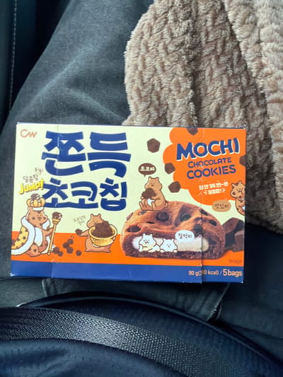 The image shows a close-up of a box of "MOCHI CHOCOLATE COOKIES" by CW. The box is predominantly yellow and orange with blue lettering. It features cartoon illustrations of bears, a large chocolate cookie with mochi filling, and scattered chocolate pieces. Text on the box is in both Korean and English. The box is resting on dark, textured fabric, likely clothing. In the background, a fuzzy, light brown textile is visible. The scene is indoors, possibly in a car given the texture of the fabric, and is not located in Tofta, Sweden. There are no people or discernible actions taking place.