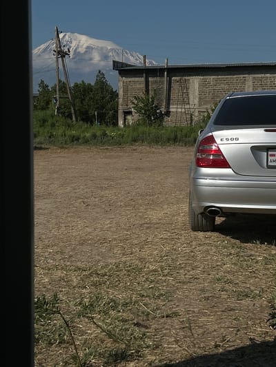 In Berkanush, Armenia, a silver Mercedes-Benz E500 is parked on a dirt and dry grass lot. The car's rear is angled towards the viewer, with its taillights and "E500" badging visible. A license plate with the letters "AM" is also present. Behind the car, a low-lying brick building with a corrugated metal roof stands under a clear blue sky. In the distance, a large, snow-capped mountain dominates the horizon, with trees and utility poles in the foreground. The scene appears to be a quiet, rural setting, possibly a residential area or a small industrial zone.