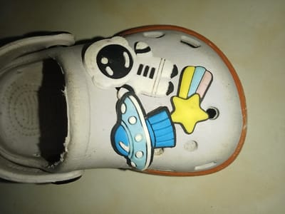 An overhead view captures a single, light grey, well-worn Crocs-style sandal oriented horizontally, with its toe facing right and the heel opening towards the left. The sandal features an orange rubber rim along its sole edge and a black heel strap secured by black rivets. The upper surface of the sandal shows minor scuffs and dirt. The internal sole is off-white and displays a circular textured pattern. Three distinct rubber charms are attached to the top of the sandal: a white astronaut with black visor details is positioned towards the upper-left, a light blue and dark blue flying saucer/UFO is located below the astronaut, and a yellow star with a rainbow-striped trail (pink, light blue, yellow) is situated to the right of the UFO. The sandal rests on a plain, light beige surface, illuminated by a direct light source. The image provides no visual data to ascertain its specific location within Umsobomvu Local Municipality, South Africa.