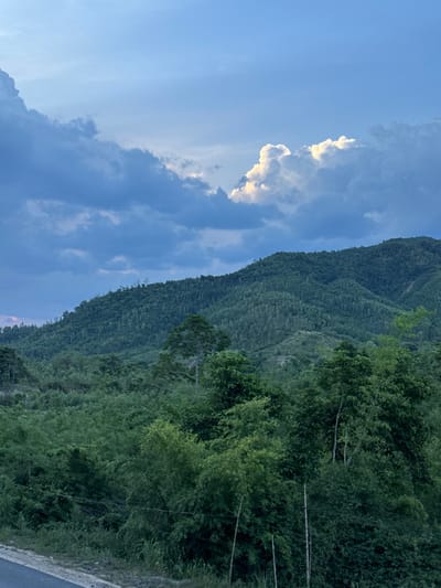 A lush green mountain range dominates the scene, covered in dense forestation that extends to the foreground. The sky above is a mix of deep blue and dark, brooding clouds, with a bright, illuminated cloud formation catching the sunlight on the right. The lower portion of the image shows a road and more vegetation, including bamboo-like plants. The lighting suggests it is either early morning or late afternoon, with the sun partially obscured by clouds. The image appears to be taken in a rural, natural setting, possibly in La Hai, Vietnam, given the dense tropical foliage and mountainous terrain. There are no people or animals visible in the image.
