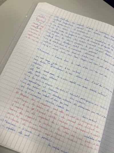 The image displays a close-up, top-down view of an open, spiral-bound notebook. The paper features French-style Séyès ruling, characterized by horizontal blue lines with faint vertical red lines forming a grid. The notebook rests on a light-colored, possibly white or off-white, flat surface. A translucent, corrugated plastic sheet, typical of a folder protector or binder, is visible along the left edge of the notebook.
The pages are filled with extensive handwritten notes primarily in blue ink, with certain words, phrases, and annotations highlighted or written in red ink. The text is in French, with structured bullet points, arrows, and circled terms. Key subjects mentioned in the notes include "culture," "régime," "inégalité sociales" (social inequalities), "Bourdieu," "Bernard Lahire," "capital économique" (economic capital), and "capital culturel" (cultural capital), indicating a focus on sociology or education. One section in red ink is circled and labeled "culture" and "régime," with arrows connecting to related concepts. No individuals are present in the scene, and no specific city location, such as Albi, France, is discernible from the visual information provided within the image frame.
