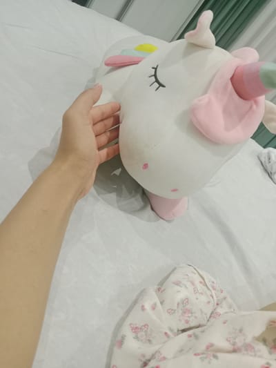 A first-person perspective shot shows a person's hand resting on a white, plush unicorn stuffed animal. The unicorn has closed eyes with embroidered eyelashes and a pink and light blue horn. Pastel-colored fabric ears and mane accents are visible. The stuffed animal is positioned on a white, creased surface, possibly a bed, with a patterned fabric in the lower right corner. The environment appears to be indoors, with hints of vertical blinds in the background. This scene is not specifically located in Keles, Uzbekistan, as no geographical markers are present.