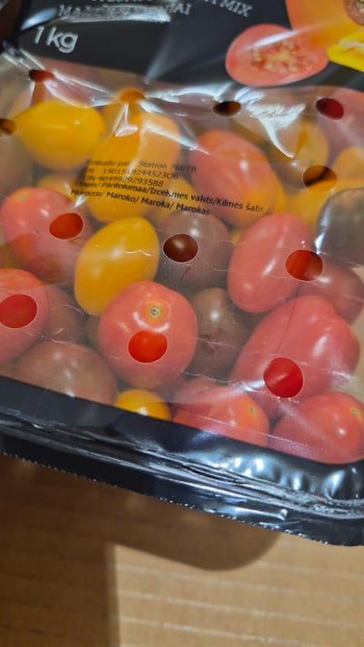The image shows a close-up view of a package of mixed cherry tomatoes, likely intended for retail sale. The tomatoes are of various colors, including red, orange, and a darker, possibly purple or brown variety. They are packed in a clear plastic container with ventilation holes. Some of the tomatoes have had portions of their skin removed or are showing signs of being cut.
Visible text on the packaging includes "MIX," "1 kg," and nutritional or origin information. Specifically, text indicates the product was "Emballé par Station 7607R" (Packaged by Station 7607R), with a lot number "150151924452306" and a GTIN "4049929293588." It also states "Origin/Päritolumaa/Izcelsmes valsts/Kilmės šalis: Morocco/Maroko/Maroka/Marokas," suggesting the tomatoes originate from Morocco. The packaging has a metallic black seal along the edge. The background appears to be a cardboard box, suggesting the product is being stored or transported. The lighting is somewhat diffused, and the focus is on the product itself. There are no people or animals visible. The overall impression is of a fresh produce item ready for consumption.