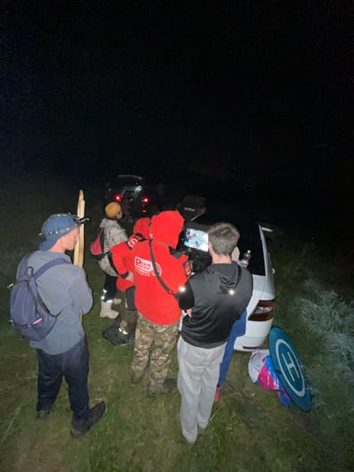 A group of at least six individuals are gathered in a grassy, outdoor area at night, near several vehicles, one of which is a white car with its trunk open. The scene appears to be in or near Badashka, Russia, given the context. Some individuals are wearing headlamps or other lighting devices. One person in the foreground wears a blue baseball cap and a dark backpack, holding a long wooden pole. Another person is dressed in a bright red hooded jacket with "PCRN RUSSIA" printed on the back. Several individuals appear to be looking at a screen, possibly a tablet or monitor, being held by one person. A blue and white helipad lies on the ground next to the white car, along with some brightly colored bags. The surrounding environment is dark, with only the lights from the vehicles and headlamps illuminating the immediate area, suggesting a search or rescue operation is underway. The ground is covered in grass and possibly some weeds.