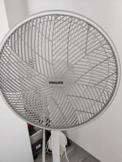 An indoor scene featuring a white Philips brand fan is captured from a close-up, head-on perspective, dominating the frame. The fan's grille is elaborate and light grey, with a repeating pattern of thin, curved bars. A prominent rectangular badge with the "PHILIPS" logo in black text is centrally located on the grille. The background is a plain, light-colored wall. The lower portion of the image reveals the fan's white stand and a hint of the floor, which appears to be tiled. In the blurred background to the left, what seem to be storage boxes or furniture are visible, some with grey and black exteriors. The scene is devoid of people or any discernible activity. The lighting is neutral and even. The image does not provide any information about the city of Grosseto, Italy.
