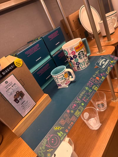 The image shows a retail display of Starbucks merchandise. Two mugs are prominently displayed on top of stacks of packaged goods. Behind the mugs is a teal sign with Korean lettering and graphics, advertising "Hangeul on the Streets Korea Edition." The sign includes references to Seoul and Korean street food. Below the sign, there is a box with a Starbucks menu sticker attached. The sticker is in Korean. There is a "Thank You!" sticker on one of the stacked boxes. The location is Gimpo-si, South Korea, based on the Korean language on the merchandise and the sign. Plates and cups are displayed on the racks behind the mugs.