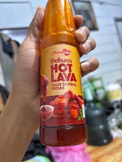A hand holds a plastic bottle of "MamaSuka Delisaos Hot Lava Saus Sambal Pedas" hot sauce. The bottle is transparent, revealing the reddish-orange sauce. The label is a light yellow color, with red and dark brown accents. The hand appears to be lightly tanned. The background is blurry, but suggests an indoor environment. The location is Mideuenjok, Indonesia.