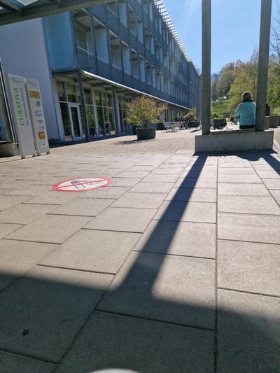 The image captures an outdoor scene at the Curativa Das Ambulante Zentrum in Rheinfelden, Switzerland. A gray, multi-storied building with numerous windows and balconies occupies the left side of the frame. In front of the building is a walkway paved with gray stones. A red and white no-smoking symbol is painted on the walkway. A white sign with the name "CURATIVA Das Ambulante Zentrum" is located near the building. A person is seated on a ledge in the background. Trees and grass are also visible in the distance, with a clear blue sky above. The image is taken during daytime, with a shadow cast across the walkway.
