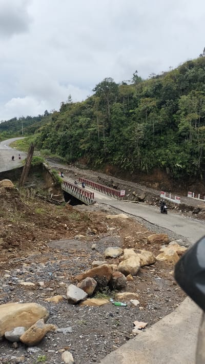 In the city of Bintangpermata, Indonesia, a damaged road is visible, with parts of the roadway collapsed. A small, red and white metal truss bridge spans a gap in the road, allowing for passage over a debris-filled channel. Several motorcycles are seen on the road, with riders navigating the damaged terrain. The surrounding environment is hilly and densely forested, with cloudy skies overhead. The foreground is filled with rocks, dirt, and some scattered debris, indicating a recent landslide or construction activity.