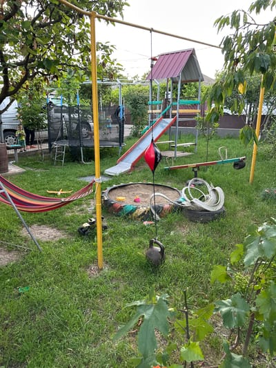 This image depicts a vibrant backyard scene in Pîrlița, Moldova, filled with various play equipment. The setting is a grassy yard with a dirt path running through it, bordered by trees and some vegetation. In the foreground, a colorful hammock with red, yellow, green, and brown stripes is suspended from a yellow metal frame, suggesting a relaxed atmosphere.
Dominating the mid-ground is a children's play structure featuring a red slide, a climbing ladder with colorful rungs, and a small house-like structure with a red roof. Nearby, a sandbox, possibly made from a repurposed tire, contains sand and some toys. A red punching bag hangs from a rope, indicating active play. A seesaw, also appearing to be made from recycled materials with a green and red frame, is positioned in the yard. A dark-colored watering can is visible on the grass.
In the background, glimpses of houses and fences suggest a residential area. There's also a trampoline with a safety net. The lighting suggests it is daytime, likely with overcast or bright skies, as there are no strong shadows. The overall impression is one of a well-used, playful outdoor space. No specific activity or event is captured, but the presence of the equipment implies children's playtime. The image is oriented sideways.