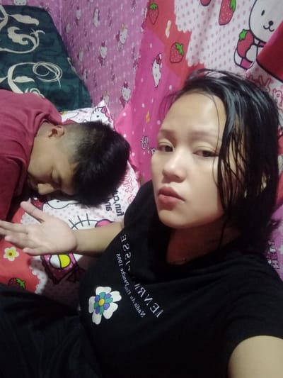 A woman poses for a selfie, lying down with her hair damp. She wears a black shirt with a flower design. Behind her, a person in a red shirt sleeps on a pillow with a Hello Kitty pattern. The bed is covered with themed bedding. The background includes a wall also decorated with Hello Kitty and strawberry motifs. The woman's expression appears somewhat exasperated.