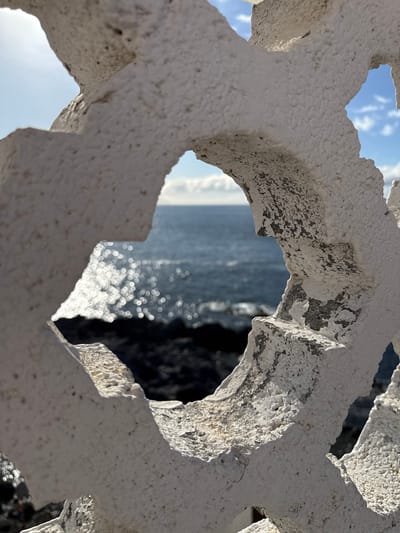 The scene is an outdoor coastal environment in Santiago del Teide, Spain. A weathered, white-painted concrete or stone decorative screen occupies the immediate foreground, exhibiting a coarse texture, extensive paint chipping, and visible surface deterioration including cracks. This screen features multiple geometric cut-out apertures resembling quatrefoils. Through these openings, a dark blue ocean extends to the horizon under a partially cloudy blue sky. The ocean surface displays numerous shimmering specular highlights from direct sunlight, creating a prominent bokeh effect. Below the water line, a dark, rugged, rocky coastline is visible. No human subjects, animals, or actions are depicted.