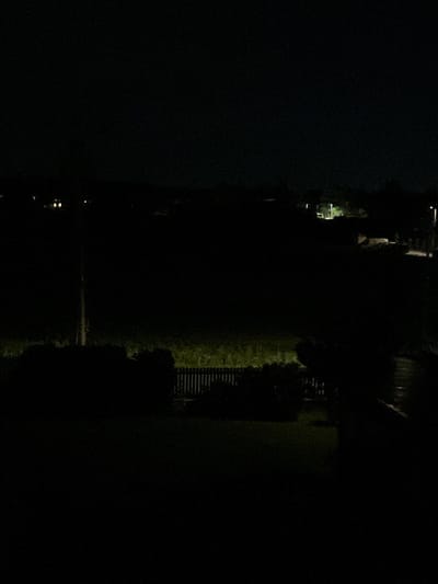 The image captures a nighttime view of a rural or suburban area, likely in Bašť, Czechia, characterized by darkness and artificial lighting. In the distance, faint illuminated structures suggest houses or buildings with interior lights on. A prominent light pole with a lamp is visible on the right side of the frame, casting a pool of light onto the ground. In the foreground, a dark fence with vertical slats is present, partially obscured by dense, dark foliage or bushes. The ground beyond the fence appears to be a grassy area illuminated by a distant light source, creating a greenish hue. The overall impression is one of a quiet, dark night with minimal activity.