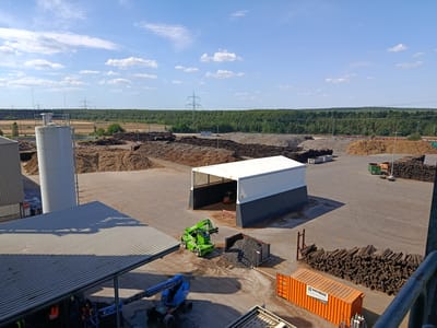 This is an aerial view of an industrial site, possibly a biomass processing facility, in Baruth/Mark, Germany. The scene is bathed in the bright, clear light of a sunny day, with scattered white clouds against a vibrant blue sky.

Dominating the foreground is a large, corrugated metal roofed structure, partially shaded. Underneath it, two individuals in high-visibility vests are working on a blue articulated boom lift. Adjacent to this structure, a green telehandler with a fork attachment is parked near a large metal skip filled with dark, granular material. A small orange forklift is visible inside a white and dark grey open-sided industrial building in the middle ground.

The yard is paved and shows tire marks. Large piles of wood chips and other biomass materials are stacked throughout the site, some appearing freshly processed and others weathered. In the background, a dense forest forms a green horizon, with power lines and transmission towers stretching across the landscape.

On the right side of the image, a bright orange shipping container with the text "HOFFMEIER" and "www.hoffmeier-berlin.de" is parked next to a neatly stacked pile of dark, cuboid-shaped material, possibly peat or compressed biomass. Another green skip is visible further in the distance.

The overall impression is one of an active industrial operation under fair weather conditions, likely during daylight hours. The presence of various machinery and the extensive piles of processed material suggest ongoing work related to resource management or energy production.