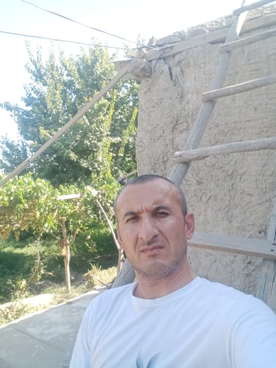 In the city of Karaganda, Uzbekistan, a man is photographed from the chest up, looking towards the camera with a neutral expression. He has short, dark hair, a receding hairline, and stubble. He is wearing a plain white t-shirt. Behind him, an aged, mud-plastered building with a wooden ladder leaning against it is visible. Lush green trees and grapevines laden with light-colored fruit are to the left of the man, suggesting an outdoor, possibly rural or semi-rural setting. The sky is clear and bright.