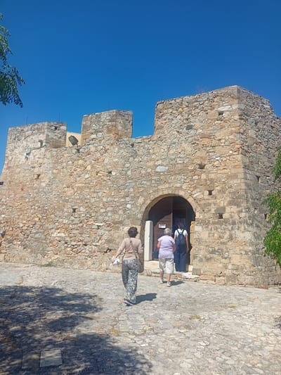 Two women and a third person are walking towards an arched entrance of a stone building, likely a historical site, under a clear blue sky. The building is constructed of rough-hewn stones and has crenellations at the top, suggesting it is a fortress or castle. The ground in front of the building is paved with cobblestones, and shadows from nearby foliage are cast across it. The woman in the foreground is wearing patterned pants and a light-colored top, while the other two individuals are entering the building through the archway. This scene is situated in the Municipal Unit of Anthidon, Greece, as indicated by the architectural style and the characteristic Mediterranean sunlight.