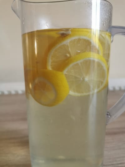 A clear plastic pitcher filled with a pale yellow liquid and several slices of lemon. The pitcher has a handle and a spout. The liquid appears to be a beverage, possibly lemonade or iced tea. The pitcher is sitting on a wooden surface. The background is out of focus and appears to be a light-colored wall. The image does not contain any information that would allow for a determination of location in Ripon, United Kingdom.