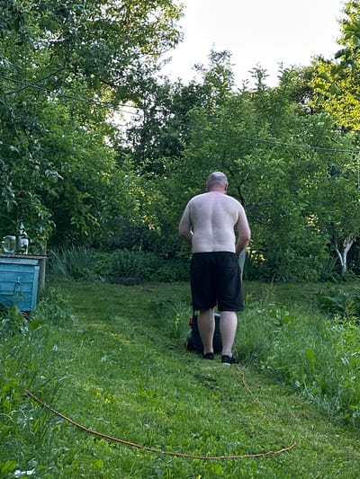 A shirtless man with short hair is mowing a grassy lawn in a backyard. He is wearing black shorts and black shoes. The man is facing away from the camera, pushing a lawnmower. The lawn is green and appears to be freshly mowed in some areas, with longer grass in others. There are several large green trees in the background, with some sunlight filtering through the leaves. To the left of the frame, there is a blue wooden structure with glass bottles on top. The location is Vydoshnia, Ukraine.