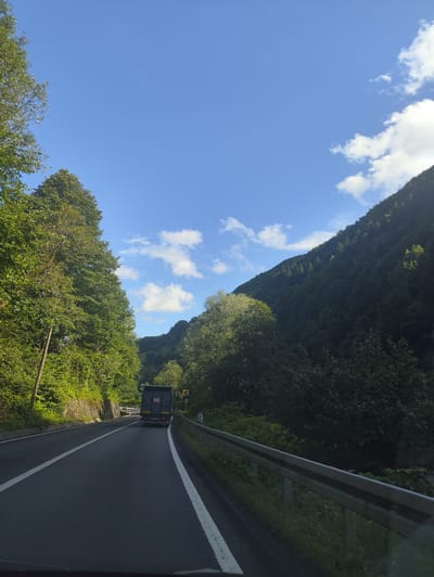 An eye-level, medium shot captures a road winding through a lush, green, mountainous landscape. The asphalt road, marked with white lines, curves to the left and recedes into the distance. A dark-colored truck is traveling away from the viewer on the road. On the left side of the road, dense trees with vibrant green foliage line a retaining wall made of stone. The right side of the road features a metal guardrail and more trees, with a steep, densely forested mountain rising on the far right. The sky above is a bright blue with scattered white clouds. The scene suggests a rural or mountainous area, likely outside of a major urban center, possibly in a region with significant forest cover like Leordina, Romania, given the context.