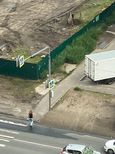 A woman with long, reddish-brown hair, wearing a dark top and light blue jeans, walks across a crosswalk on a paved road. She is heading away from the viewer. To the right of the road, there is a sidewalk adjacent to a green fence. Beyond the fence is a construction site with muddy ground, tire tracks, and tall green grass. A white truck with a closed trailer is parked on the paved area near the sidewalk. Traffic lights and pedestrian crossing signs are visible on a pole near the fence. In the background, other vehicles and a glimpse of buildings are present. The scene appears to be in an urban or suburban area.