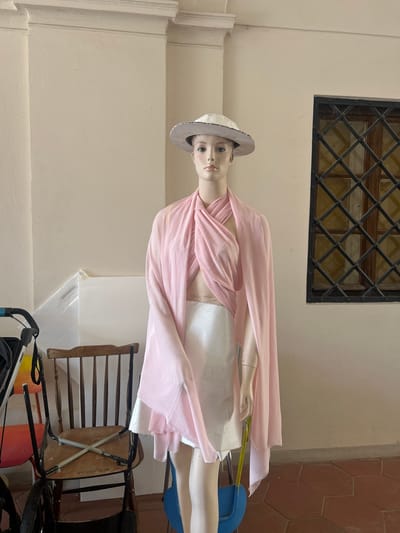 A mannequin is dressed in a pink wrap and a white skirt, wearing a grey hat. It is positioned in front of a cream-colored wall with a barred window above. To the right, there is a wooden chair and what appears to be a stroller. The lighting suggests it is daytime. The setting is outdoors or in a covered outdoor space, possibly a courtyard or porch, given the tile flooring visible on the right.