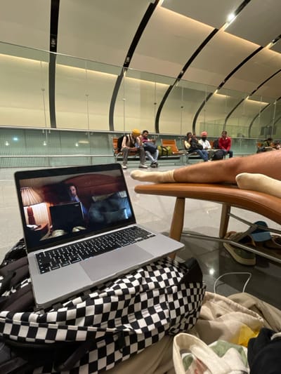 This image depicts an interior scene within a modern transportation terminal, located in Seeb Province, Oman, likely Muscat International Airport. In the foreground, a silver MacBook Pro laptop is open, displaying a video of a bearded man seated at a desk with a laptop and a table lamp, resting on a black and white checkered backpack. To the right, a person's bare leg with visible hair, wearing a white sock on the left foot, is extended and rests on a brown wooden lounge chair with metal supports. A second white-socked foot is partially visible on the same chair. Below the chair, two pairs of casual shoes—one light blue and brown, one olive green and brown—are visible on the polished dark floor, alongside various folded textiles in beige, yellow, green, white, and black.

In the midground, behind a glass partition, multiple individuals are seated on orange and brown modular airport benches. From left to right, identifiable figures include a man in a yellow turban looking down, a man in a dark shirt, a man in a dark purple shirt, a woman in a dark outfit, a man in a white turban, and a man in a red jacket. All appear to be waiting. The background features contemporary airport architecture: a high, curved, light-colored ceiling with dark structural beams, and a patterned wall behind the seating area. Artificial lighting illuminates the spacious, clean environment.