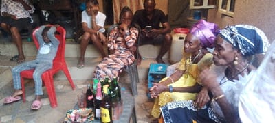 A group of people is gathered outdoors. A child in pajamas sits on a red plastic chair. An adult in a patterned dress sits on a metal chair. Two older women wearing head wraps are seated, and an older man is looking at his phone. Bottles and snacks are on the ground. There are also two white plastic water jugs and a small blue machine.