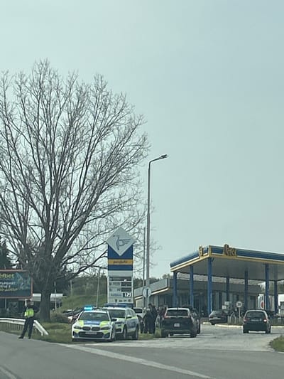 In Tserovo, Bulgaria, a service station displays a price board showing fuel costs. A police vehicle with flashing blue lights is positioned nearby. Bare trees frame the scene. Several other vehicles are present, parked near the station's gas pumps. A billboard is visible near the road. A uniformed figure stands near the billboard. The overall environment appears overcast.